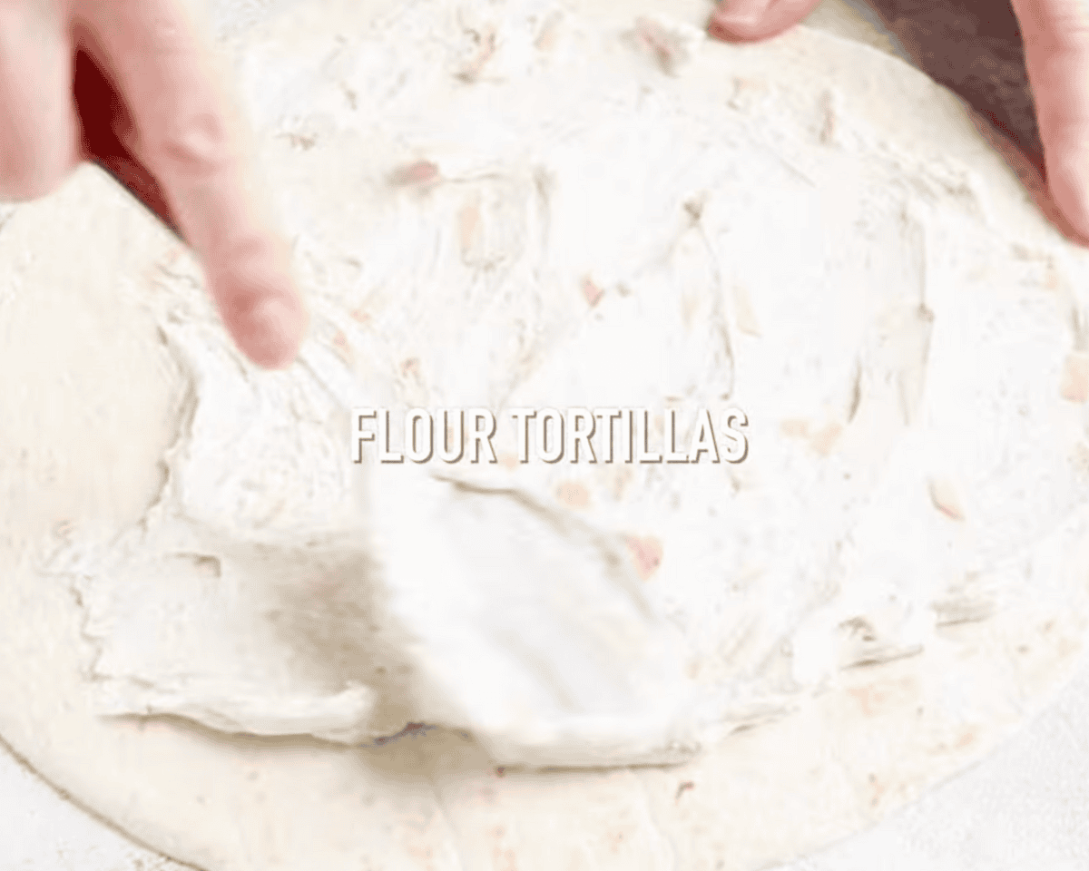 A flour tortilla being spread with a creamy cheese mixture using a spatula, with overlaid text reading "FLOUR TORTILLAS."