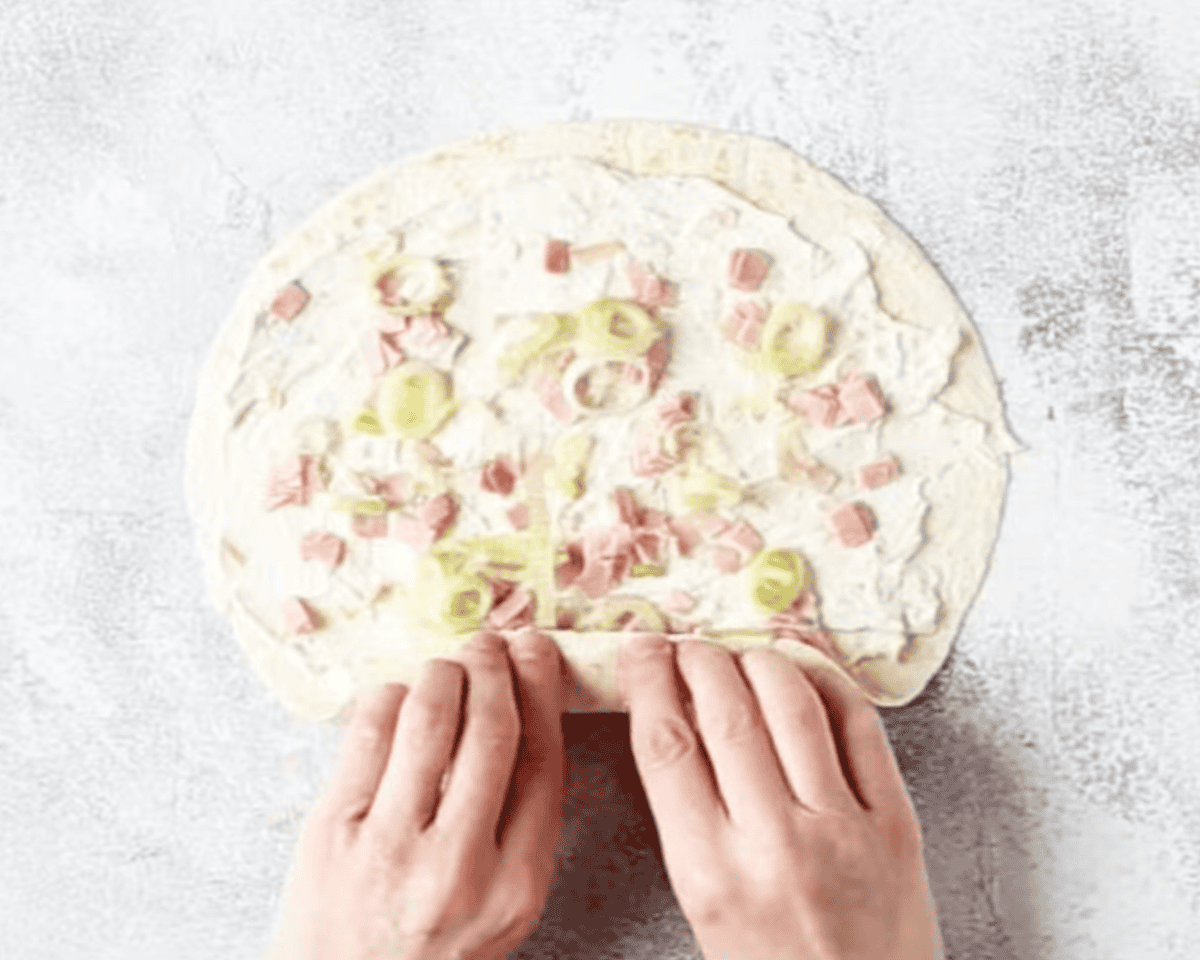 Hands rolling up a tortilla filled with a creamy spread, diced ham, and sliced green onions on a light, textured surface.