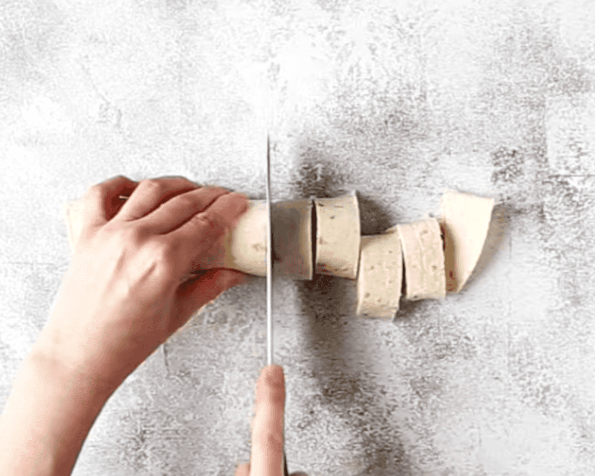  A pair of hands slicing a rolled-up tortilla into even pinwheels on a textured surface. The pinwheels reveal a creamy filling with visible ingredients inside.