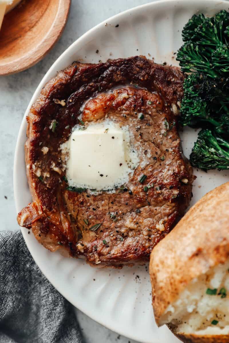 close up of a pan seared ribeye on a white plate with butter on top and a side of baked potato and broccoli rabe.