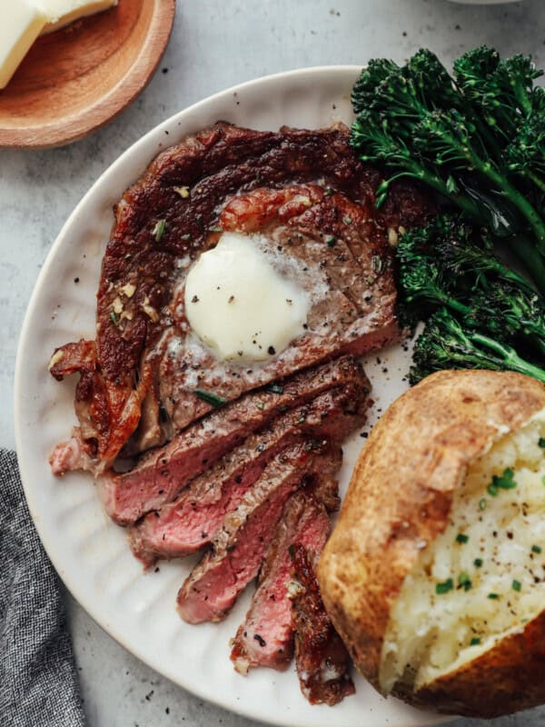 overhead view of a sliced pan seared ribeye on a white plate topped with butter and a side of baked potato and broccoli rabe.