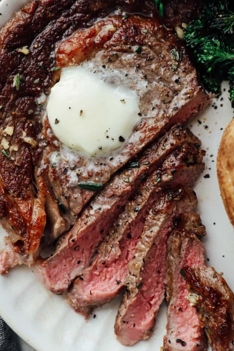 close up view of a sliced pan seared ribeye on a white plate with butter on top.