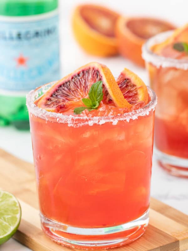 close up three-quarters view of a blood orange margarita on a wooden cutting board garnished with a blood orange slice and mint leaves.