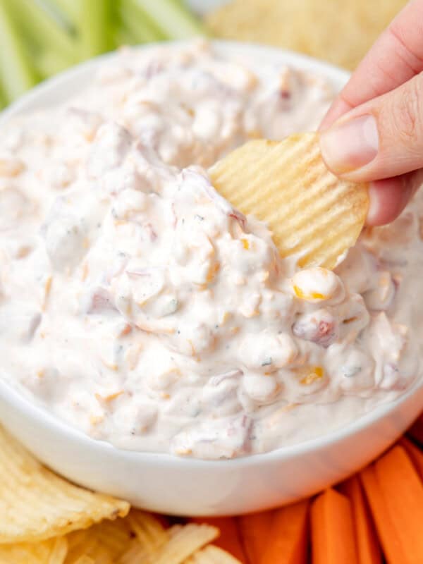 three-quarters view of a hand dipping a wavy potato chip into fiesta ranch dip in a white bowl surrounded by wavy potato chips, carrot sticks, tortilla chips, and celery sticks.