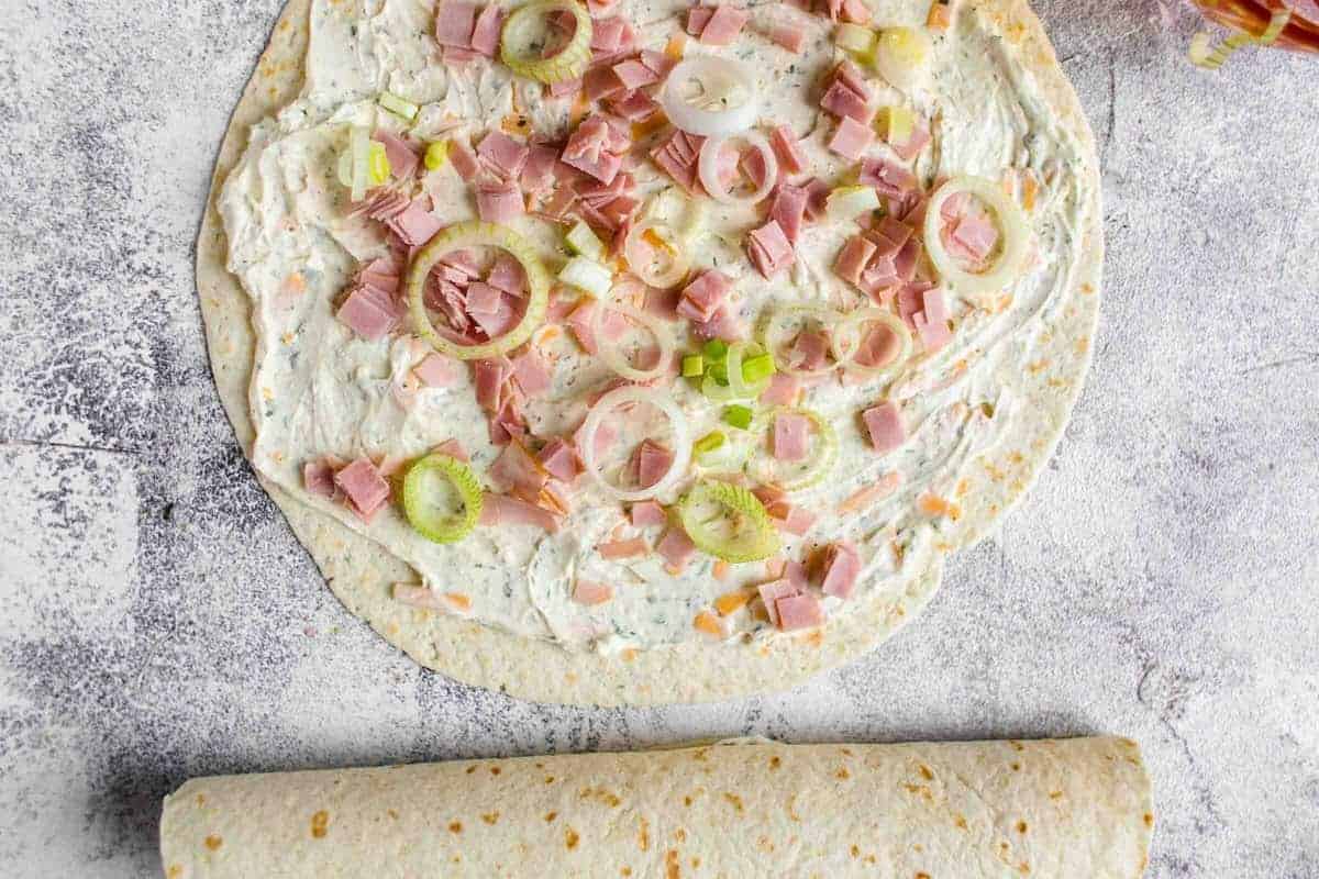 A fully assembled tortilla laid flat with a creamy cheese spread, diced ham, and sliced green onions, with another rolled tortilla at the bottom of the frame.