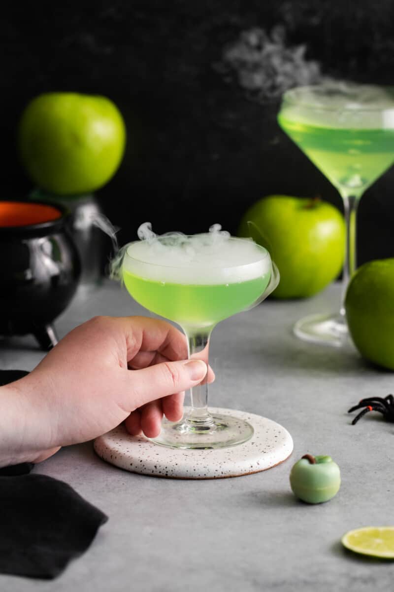 a hand grabbing a smoking poison apple martini on a white coaster.