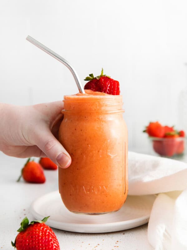 a hand grabbing a strawberry mango smoothie in a mason jar with a stainless steel straw.