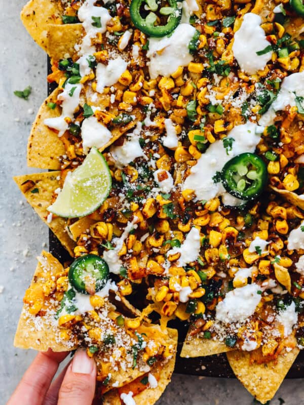 a hand grabbing from the corner of a baking sheet filled with street corn nachos.