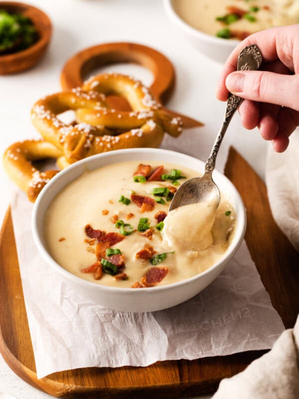 three-quarters view of a hand dipping a spoon into a serving of beer cheese soup in a white bowl on a cutting board with 2 soft pretzels.