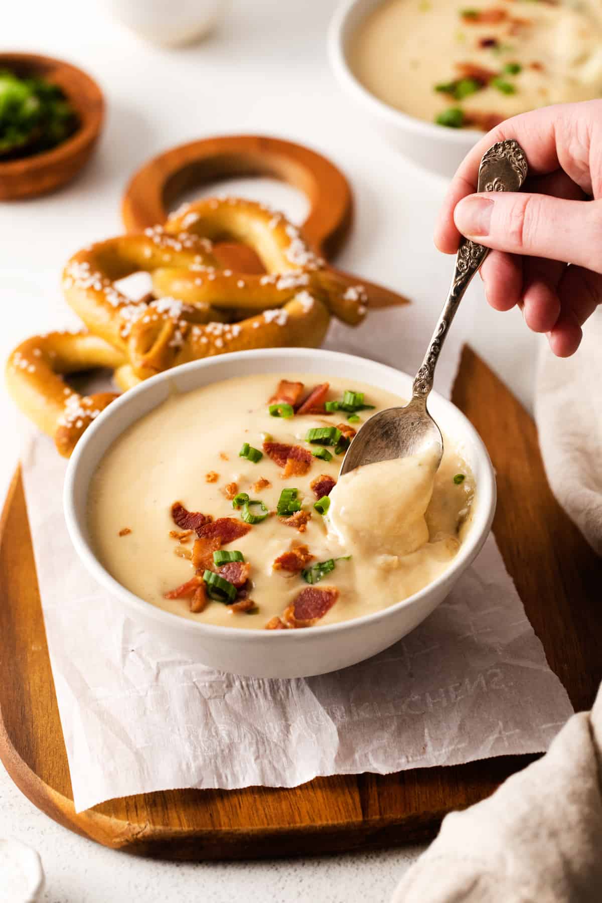 three-quarters view of a hand dipping a spoon into a serving of white cheddar beer cheese soup in a white bowl on a cutting board with 2 soft pretzels.