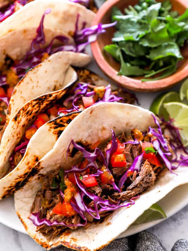 overhead view of 4 barbacoa tacos on a white plate with a bowl of cilantro.