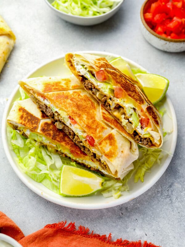 three-quarters view of a stacked halved crunchwrap supreme on a white plate with lettuce and lime wedges.