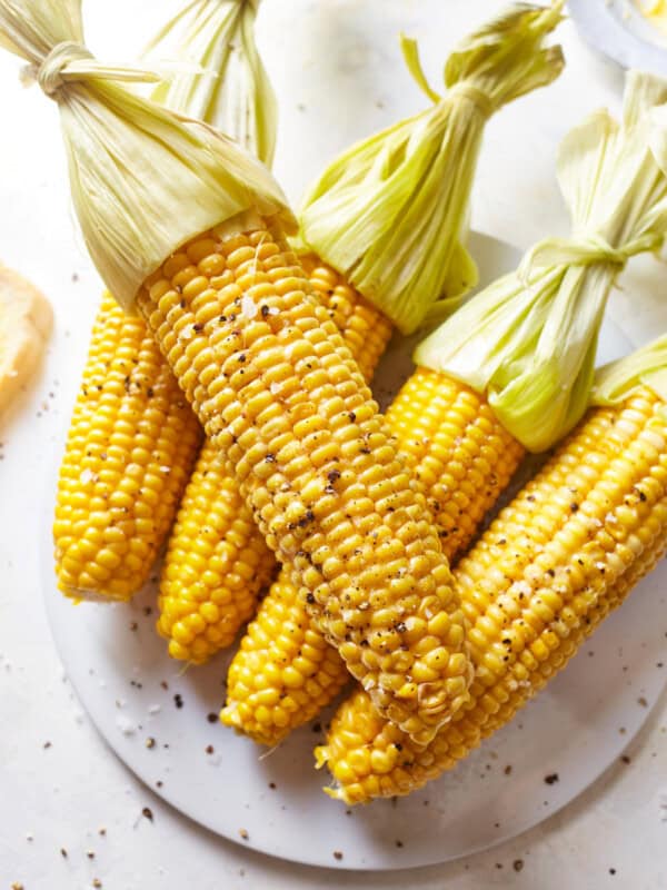 5 ears of corn on the cob with their husks tied back on a glass plate.