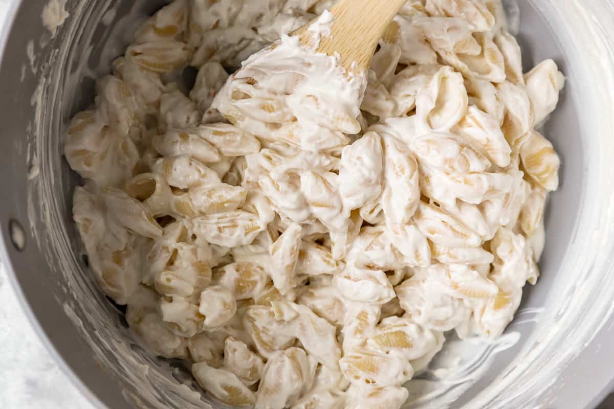 shell pasta mixed with cream cheese in a white bowl with a wooden spoon.