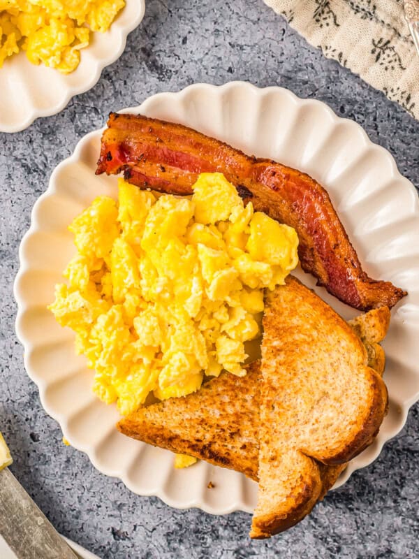 overhead view of scrambled eggs on a white plate with toast and bacon.