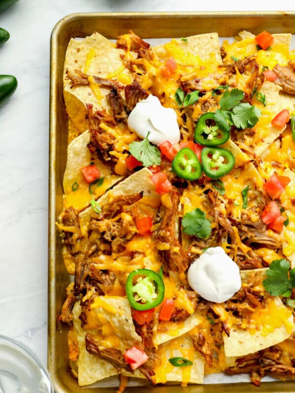 partial overhead view of pulled pork nachos on a baking sheet topped with sour cream, jalapenos, tomatoes, and cilantro.