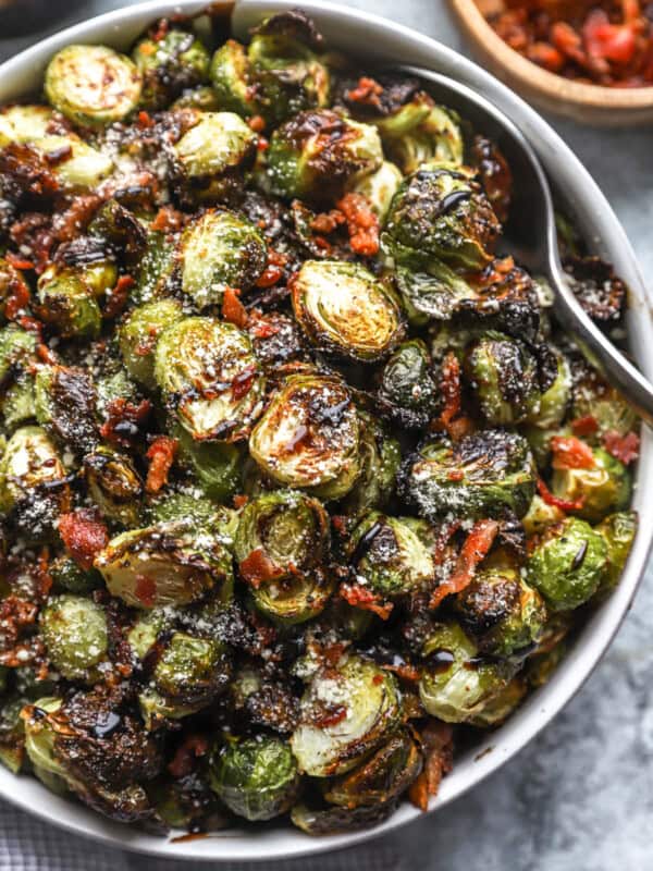 partial overhead view of roasted brussels sprouts topped with bacon, cheese, and balsamic in a serving bowl with a serving spoon.