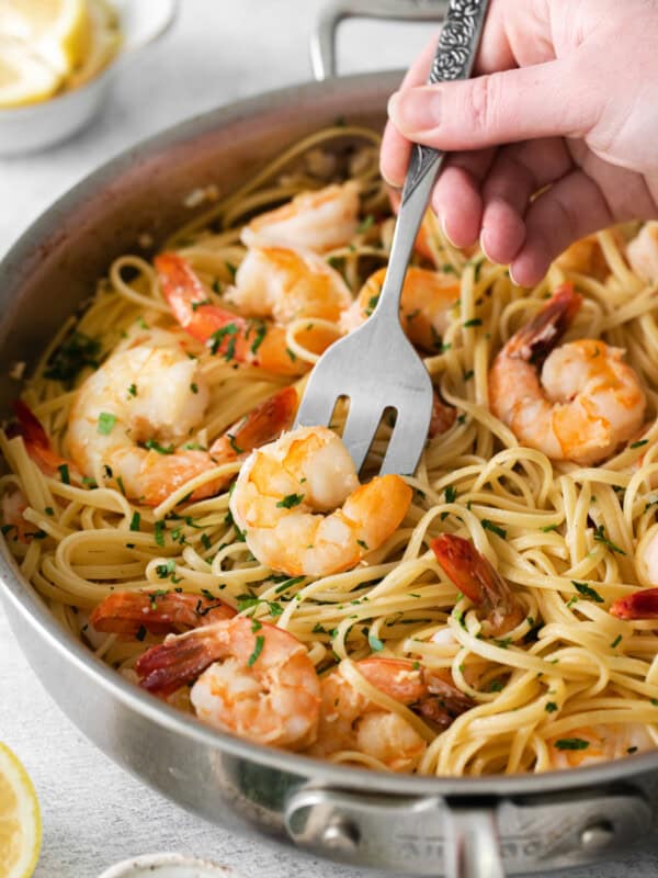 a hand using a slotted spoon to scoop a shrimp from a bed of pasta in a frying pan.