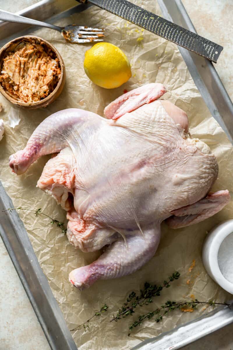 raw spatchcock chicken on a parchment-lined baking sheet surrounded by spice rub ingredients.