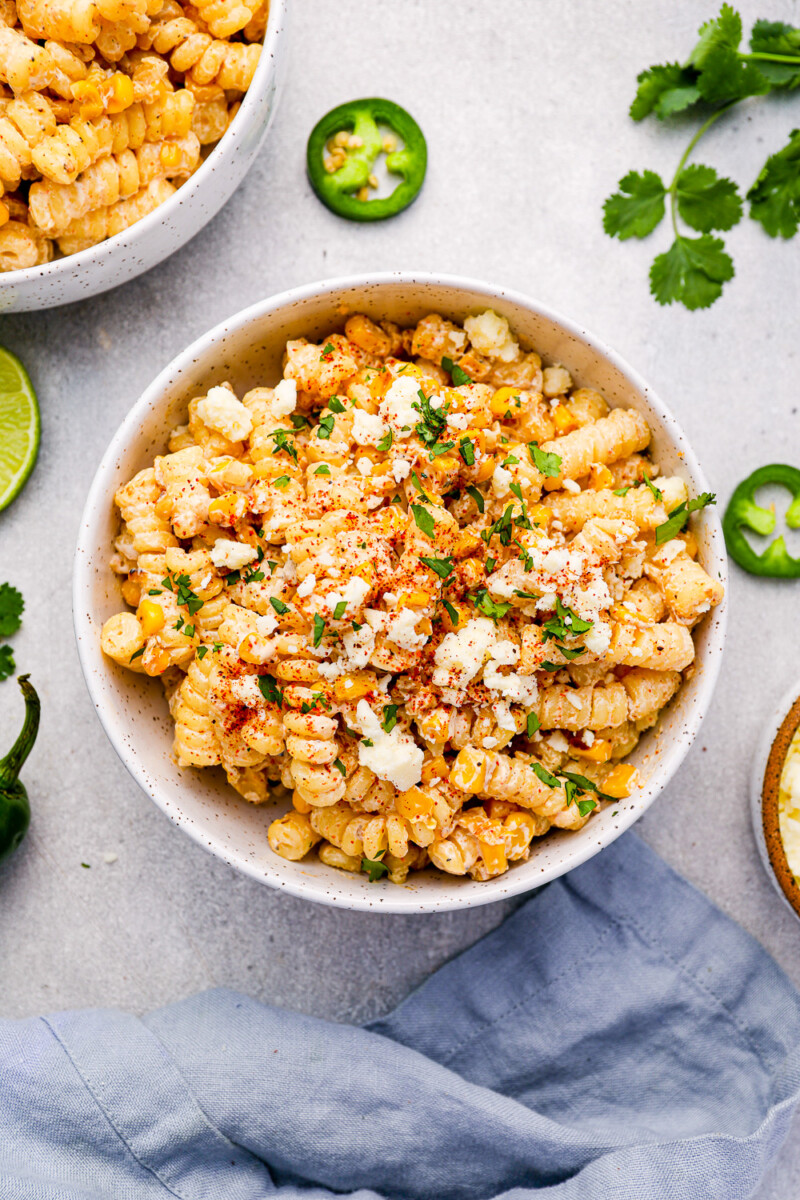 overhead view of a serving of street corn pasta salad in a white bowl surrounded by sliced jalapeños.