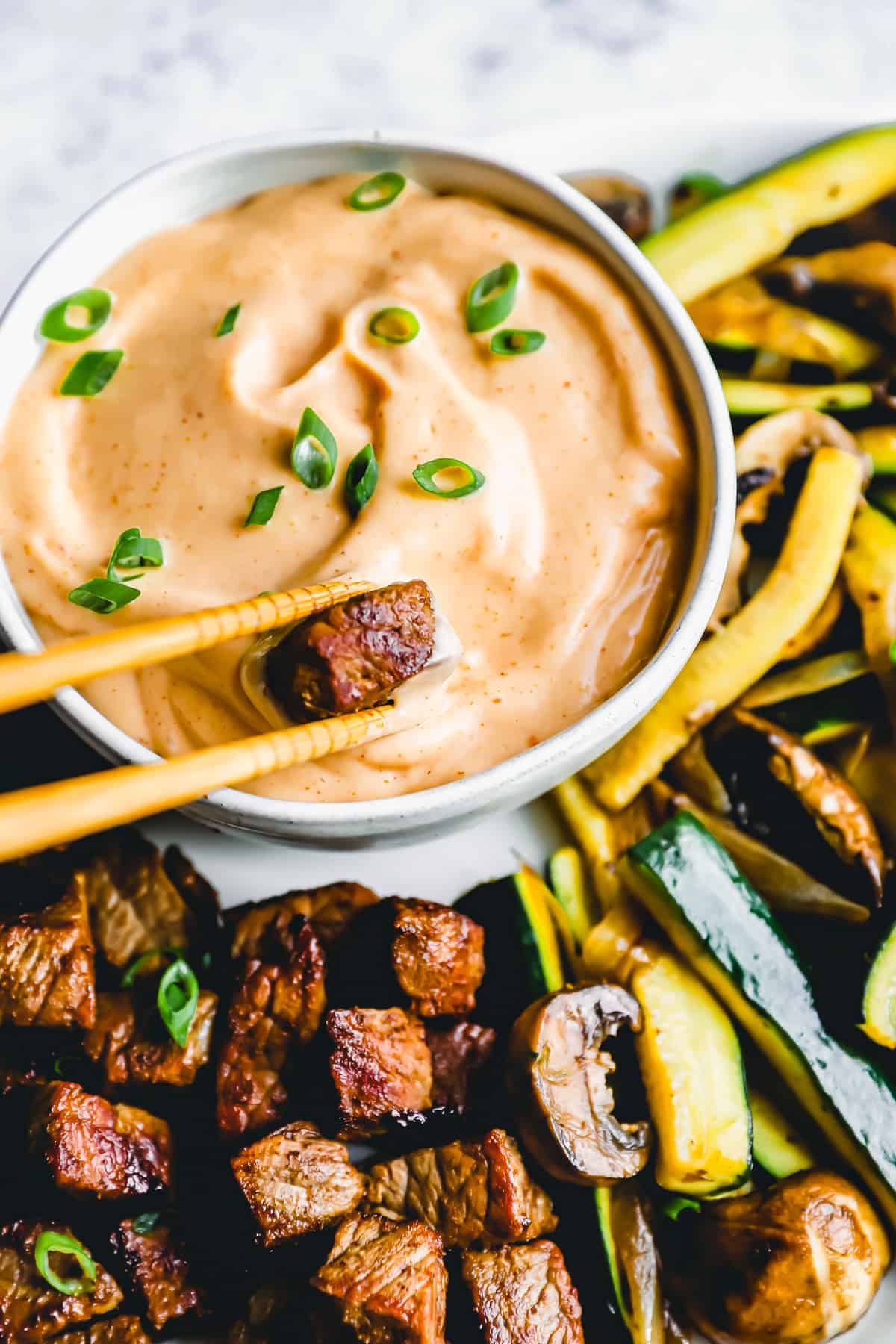 three-quarters view of a pair of chopsticks dipping a cube of steak into DIY yum yum sauce in a white bowl.