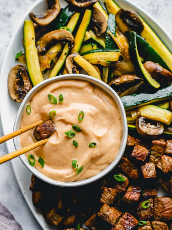 overhead view of a pair of chopsticks dipping a cube of steak into yum yum sauce in a white bowl.