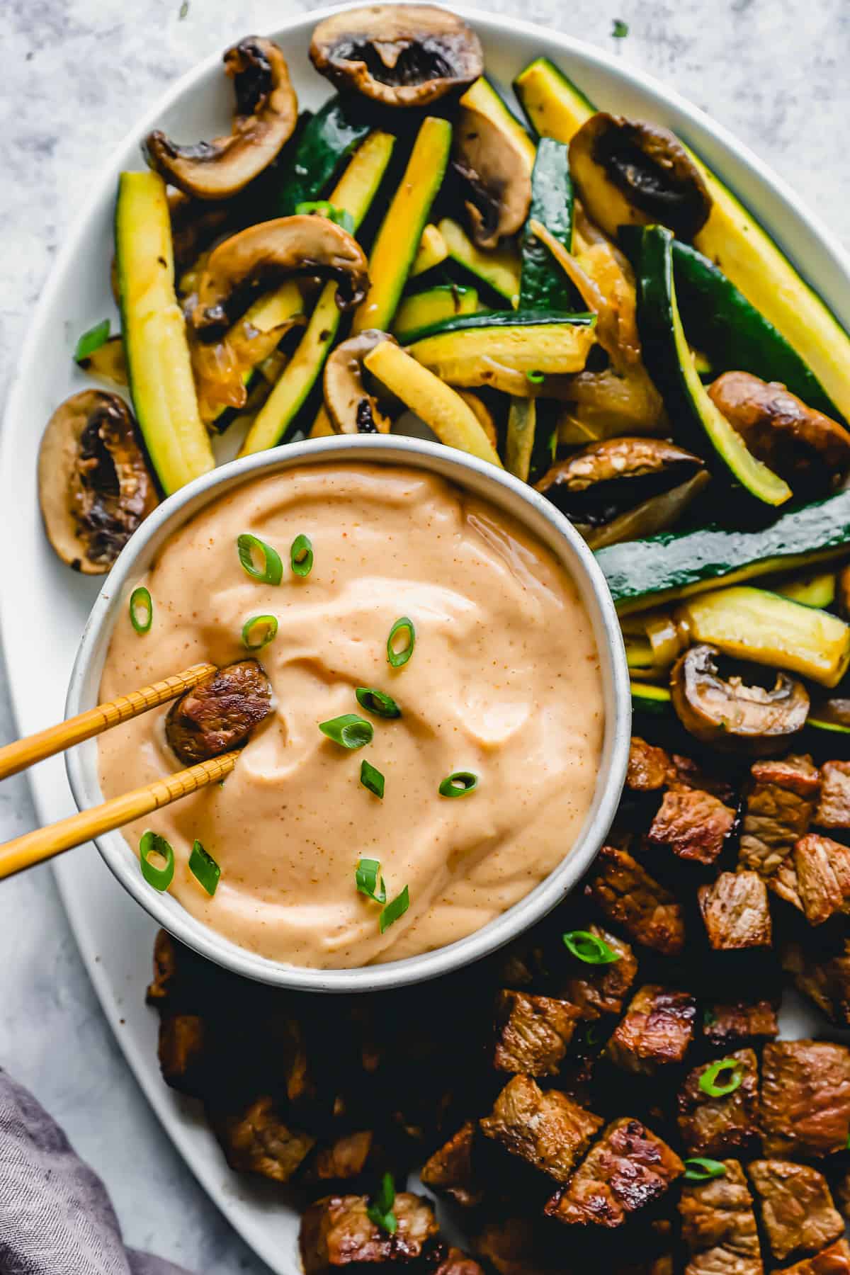 overhead view of a pair of chopsticks dipping a cube of steak into yum yum sauce in a white bowl.