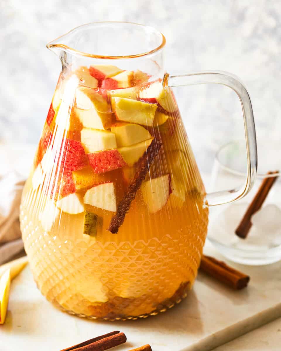 a glass pitcher full of caramel apple sangria, with chopped apples and cinnamon sticks visible in the liquid