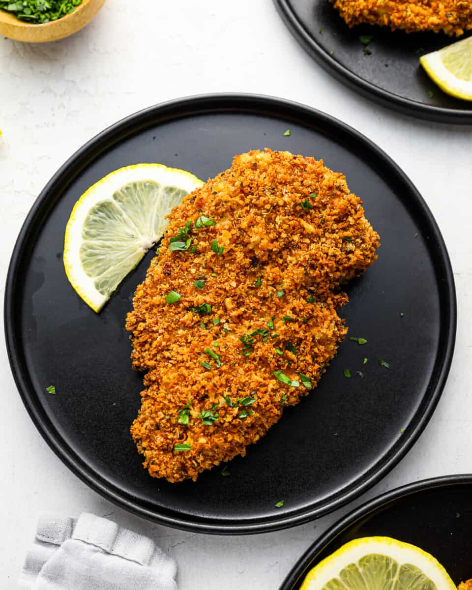 a panko crusted chicken breast on a black plate with a lemon slice.