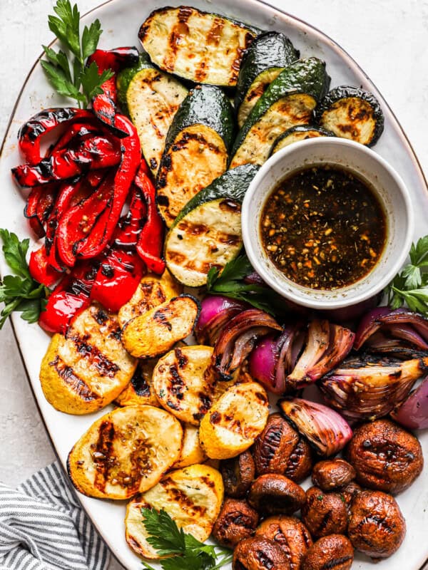 overhead view of grilled vegetables on a white oval plate with marinade in a small bowl.