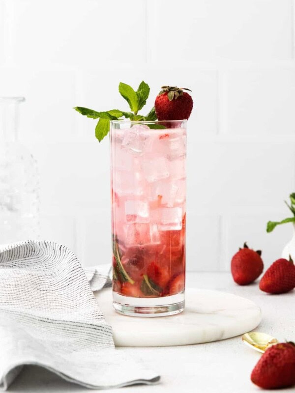 side view of a strawberry mojito in a highball glass on a white plate topped with mint sprigs and a strawberry.