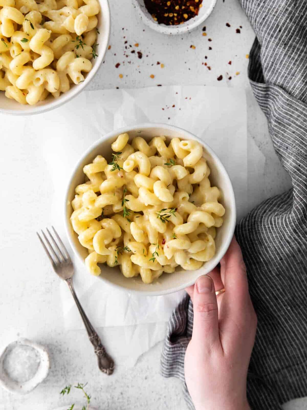 overhead view of a hand holding a single serving of white cheddar mac and cheese in a white bowl with a fork on the side.