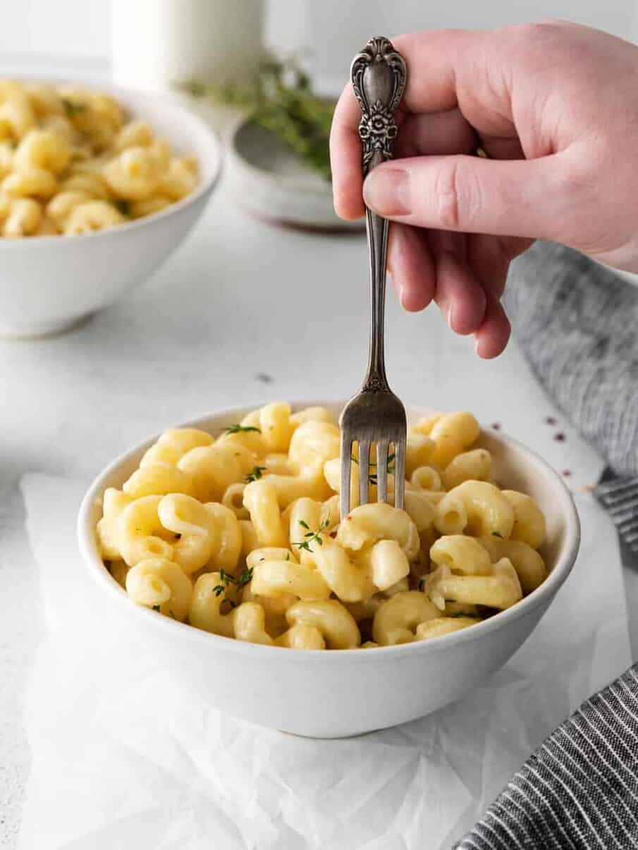 three-quarters view of a hand stabbing a fork into a serving of white cheddar mac and cheese in a white bowl.