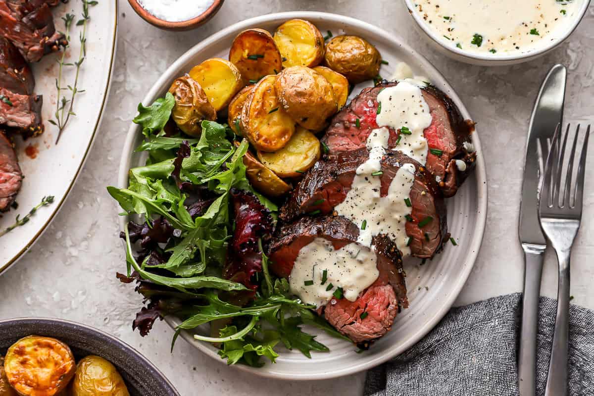 overhead view of slices of beef tenderloin on a white plate topped with cream sauce and a side of salad and potatoes.