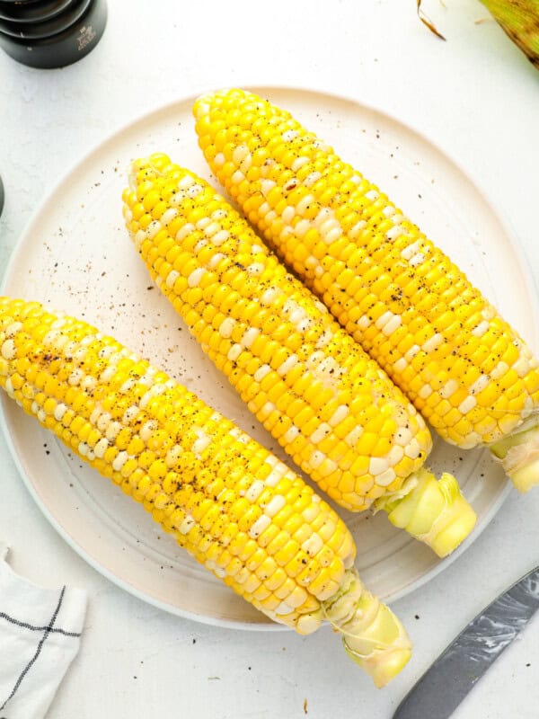 overhead view of 3 grilled corn cobs on a white plate seasoned with salt and pepper.