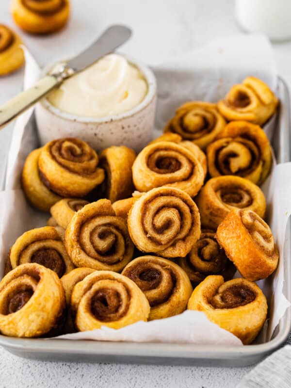 three-quarters view of mini cinnamon rolls in a serving tray with a small pot of frosting and a knife.