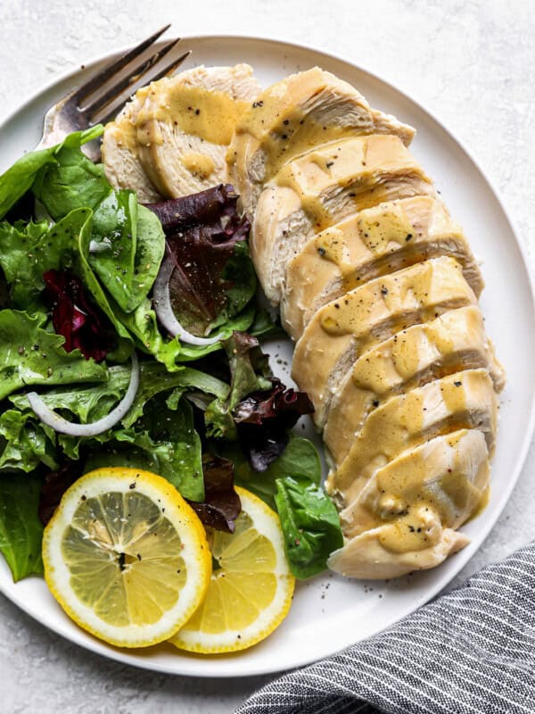 overhead view of a sliced poached chicken breast on a white plate with greens, lemon slices, and a fork.