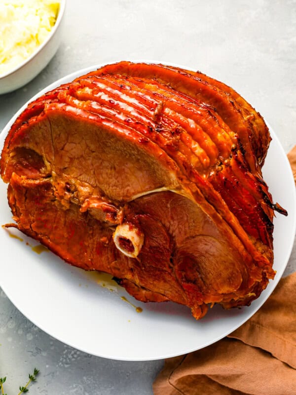 A roasted ham on a plate next to a bowl of mashed potatoes.