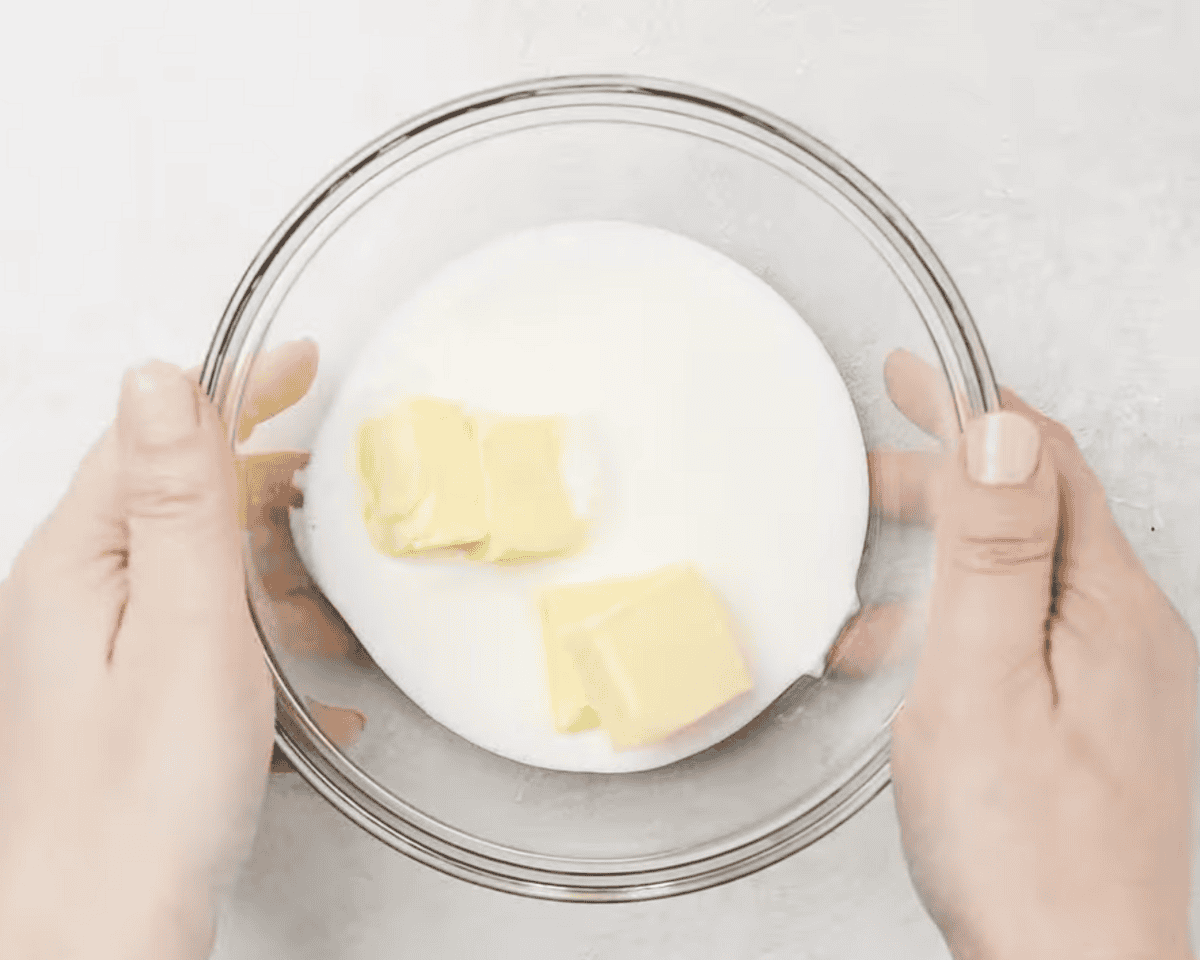 A person holding a clear glass bowl filled with buttermilk and chunks of butter, preparing to warm or mix the ingredients.