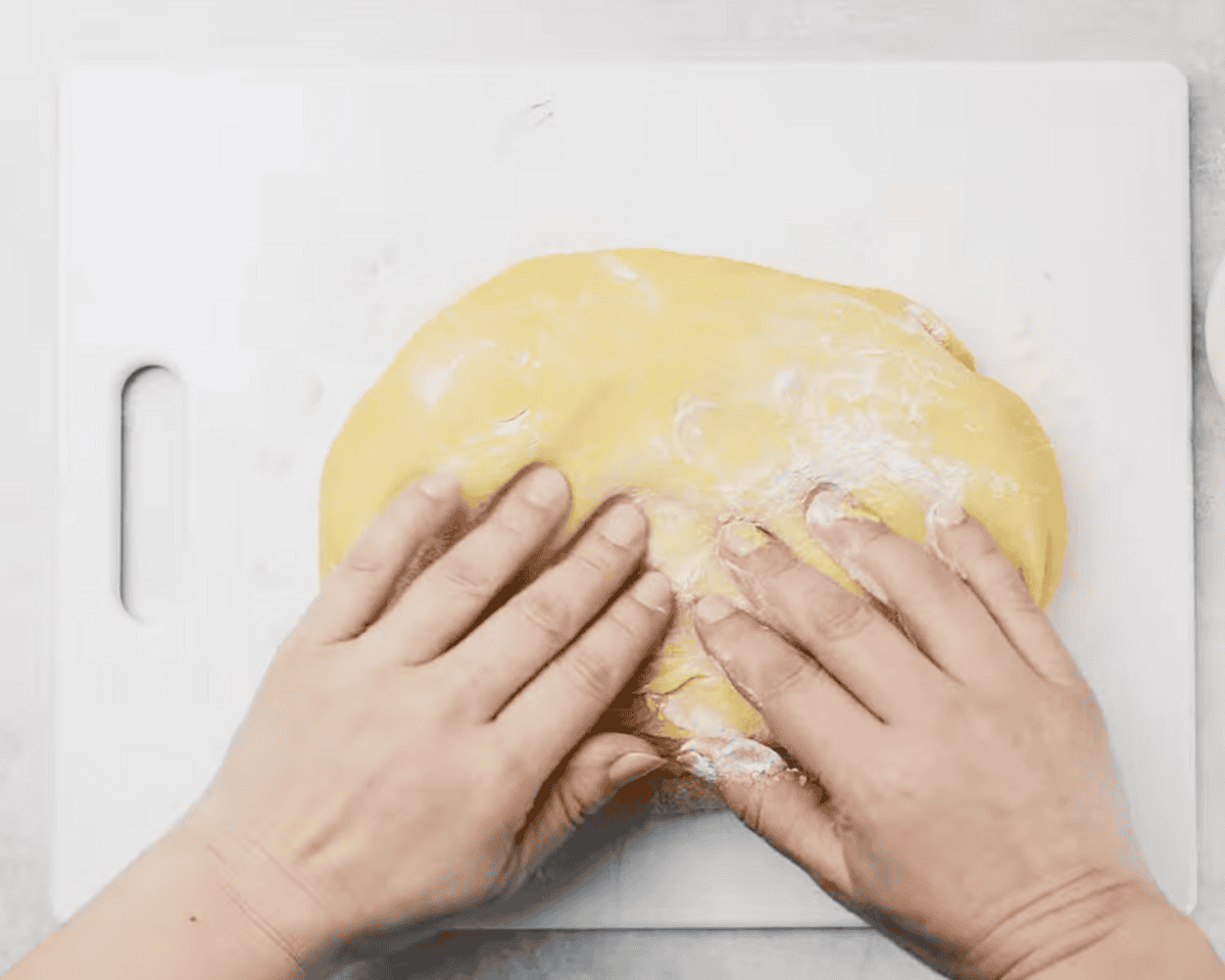 Two hands kneading a soft dough on a white cutting board, with a small bowl of flour nearby.