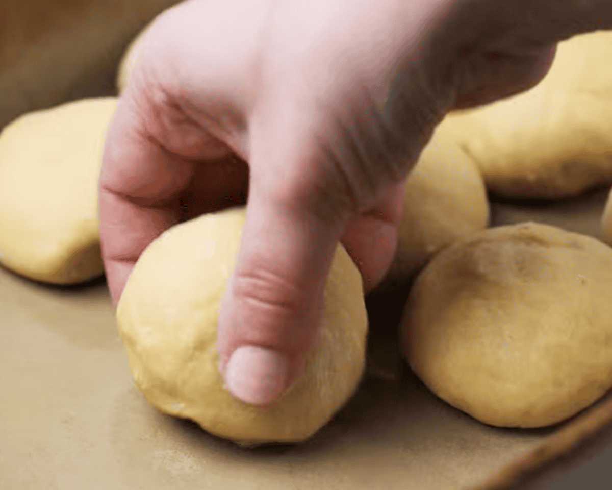 A close-up of a hand carefully placing a dough ball into a baking dish with other shaped rolls.