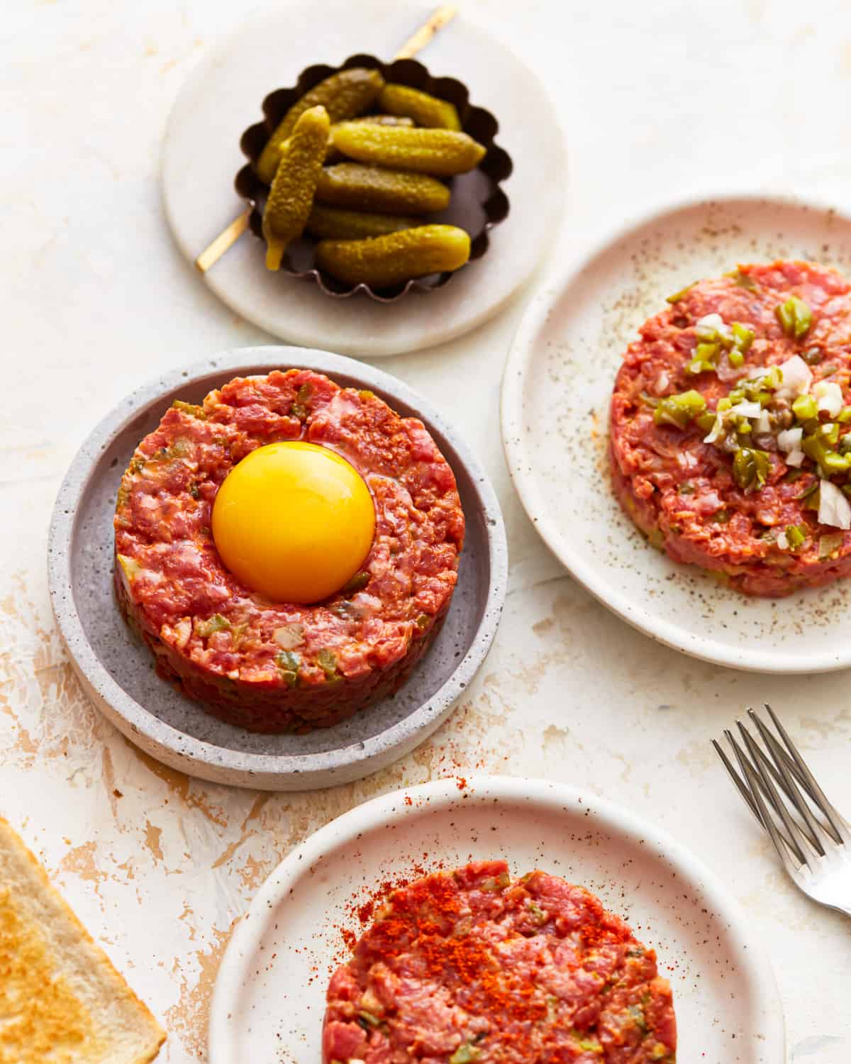 Three plates with beef tartare on them, one topped with paprika, one topped with egg, and one topped with minced pickles.