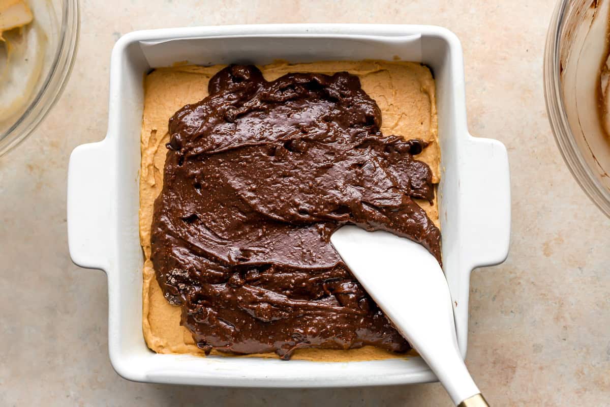 brownie batter spread over pumpkin filling over brownie batter in a square baking dish.