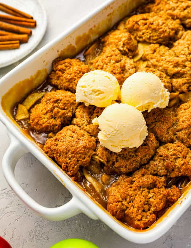 Apple cobbler in a baking dish with ice cream and cinnamon sticks.
