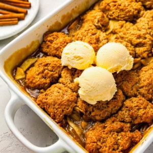 Apple cobbler in a baking dish with ice cream and cinnamon sticks.