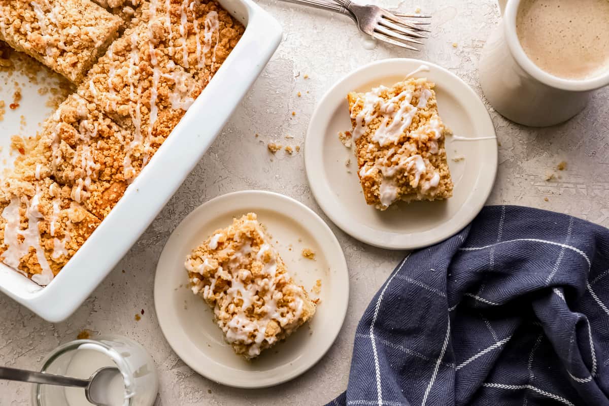 Pieces of apple cinnamon coffee cake on white plates, on a table top with forks, mugs of coffee, and a dish towel scattered around.