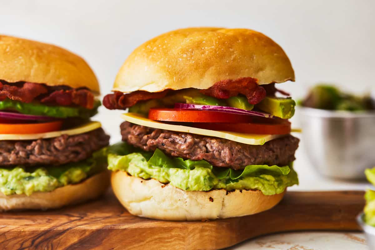 Two avocado bacon burgers are sitting on a wooden cutting board.