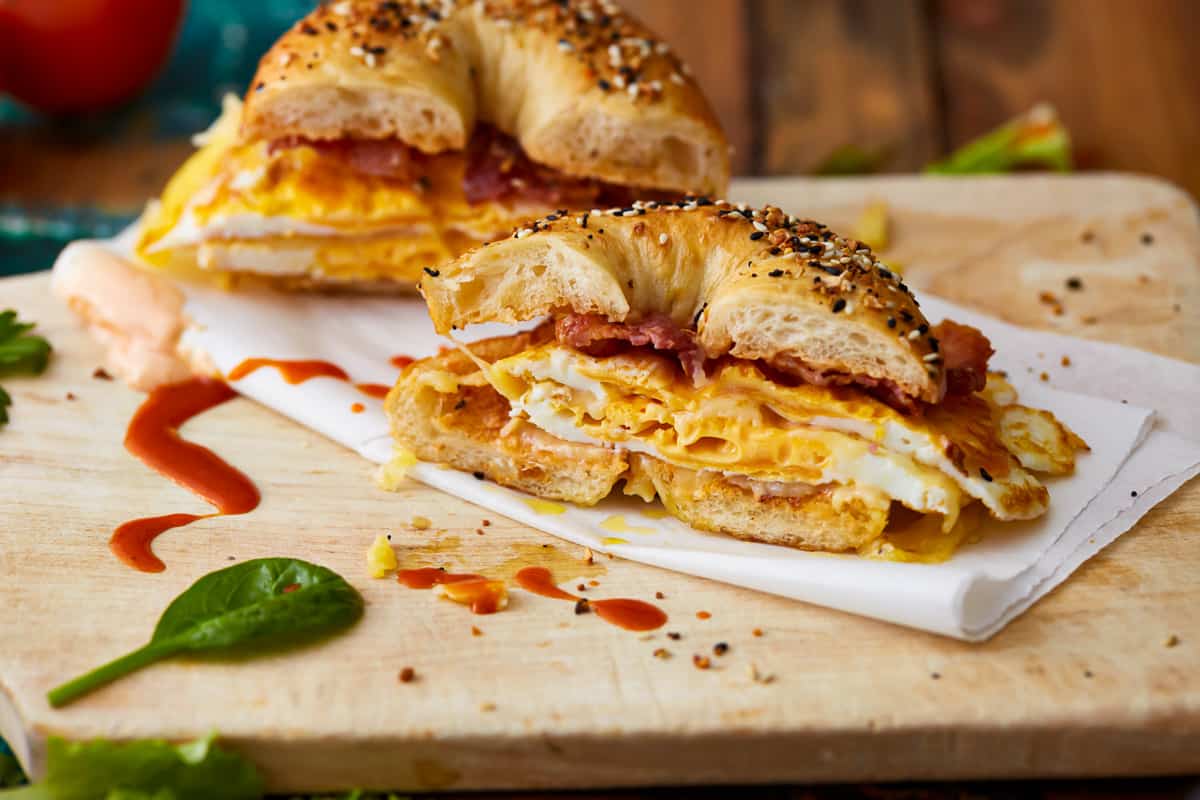 A bacon egg and cheese bagel cut in half, on a cutting board with hot sauce.