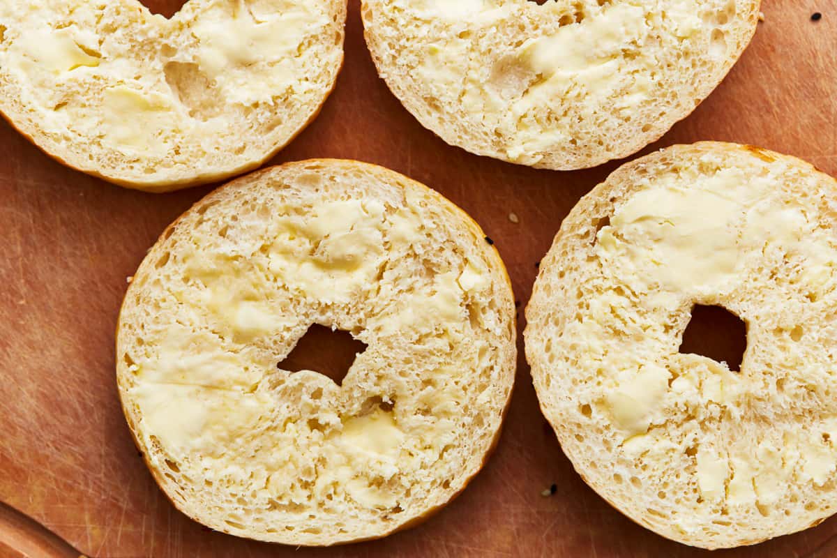 Four buttered bagels on a wooden cutting board.