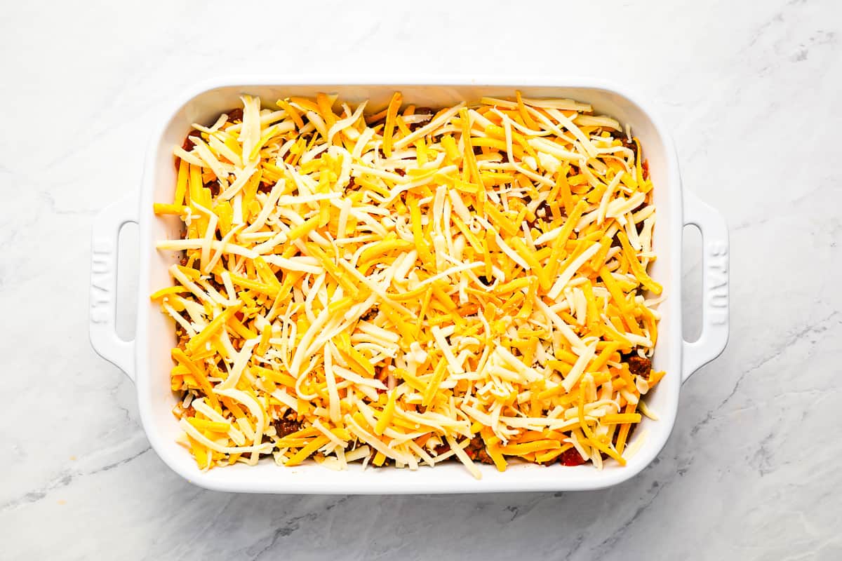 Taco dip topped with shredded cheese, in a white casserole dish.